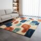 A contemporary hand-tufted rug featuring large, organic pebble shapes in terracotta orange, sapphire blue, and seafoam green on a cream background, styled in a minimalist living room with a grey sofa and large window.