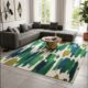 Modern living room featuring a large hand-tufted abstract area rug with vibrant green, teal, and navy blue brushstroke patterns on a cream base, styled under a gold coffee table and grey sectional sofa.