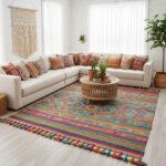 A colorful bohemian tribal rug with teal, orange, and pink horizontal geometric patterns and multi-colored tassels, styled in a bright white living room with a beige sectional sofa and macramé wall hanging.